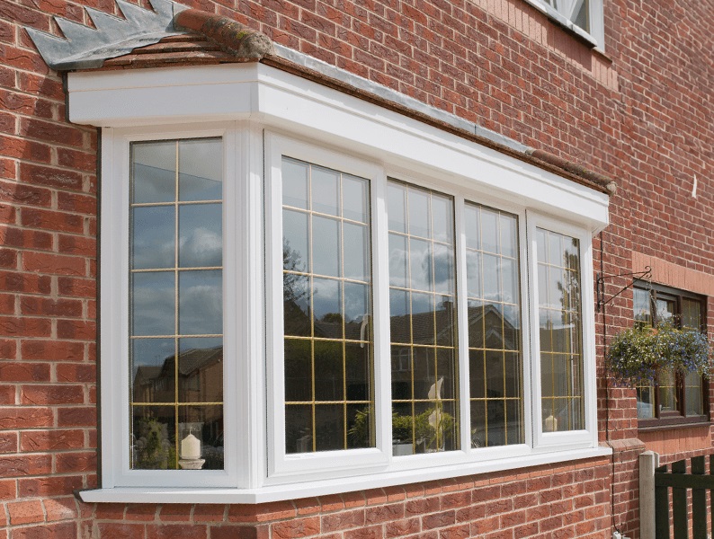 A white uPVC bay window with grid designs installed in a red brick house, reflecting a clear sky and the surrounding greenery, with a hanging flower basket adding a touch of charm to the quaint exterior.