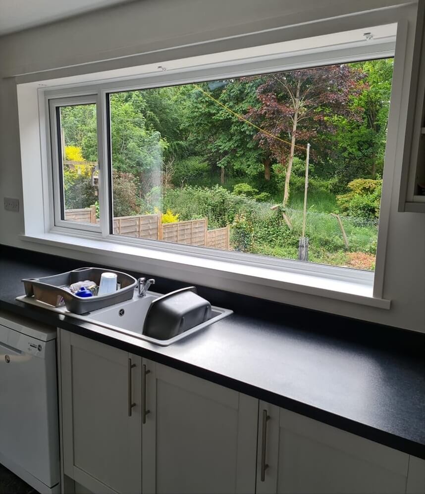 The image shows a kitchen interior with a large window above the sink, offering a view of a garden. The window appears to be a white uPVC casement type, with one side openable. The kitchen has white cabinets and a dark countertop, and there's a dish rack with a few dishes in it on the sink. The view outside includes a lush green garden with a variety of trees and shrubs, and a fence dividing the property.